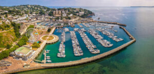 Aerial view of Torquay Marina on a sunny day, featuring yachts, boats, promenade, and blue sea along the English Riviera coastline